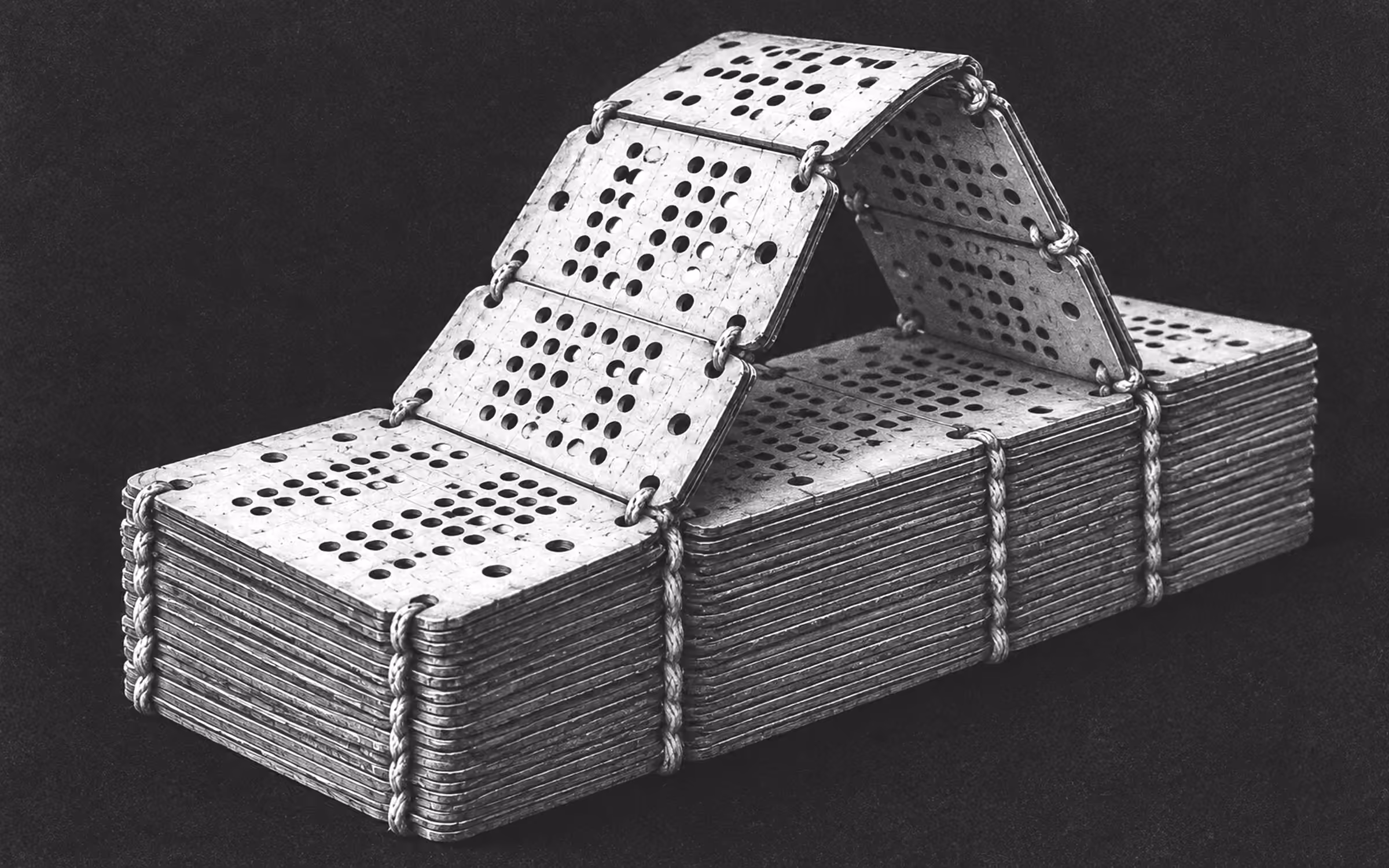 A dense stack of laced Jacquard punch cards on a dark field, with a short linked length of cards lifted from the stack and folded back in a low arch.